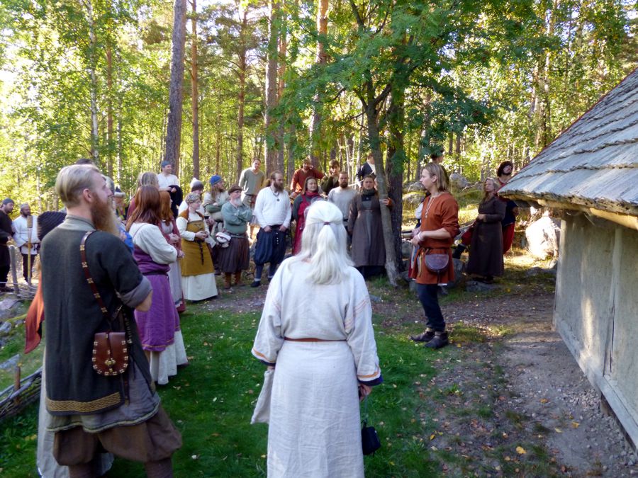 Personer som sitter i gräset och lyssnar på en guide som står framför ett hus.